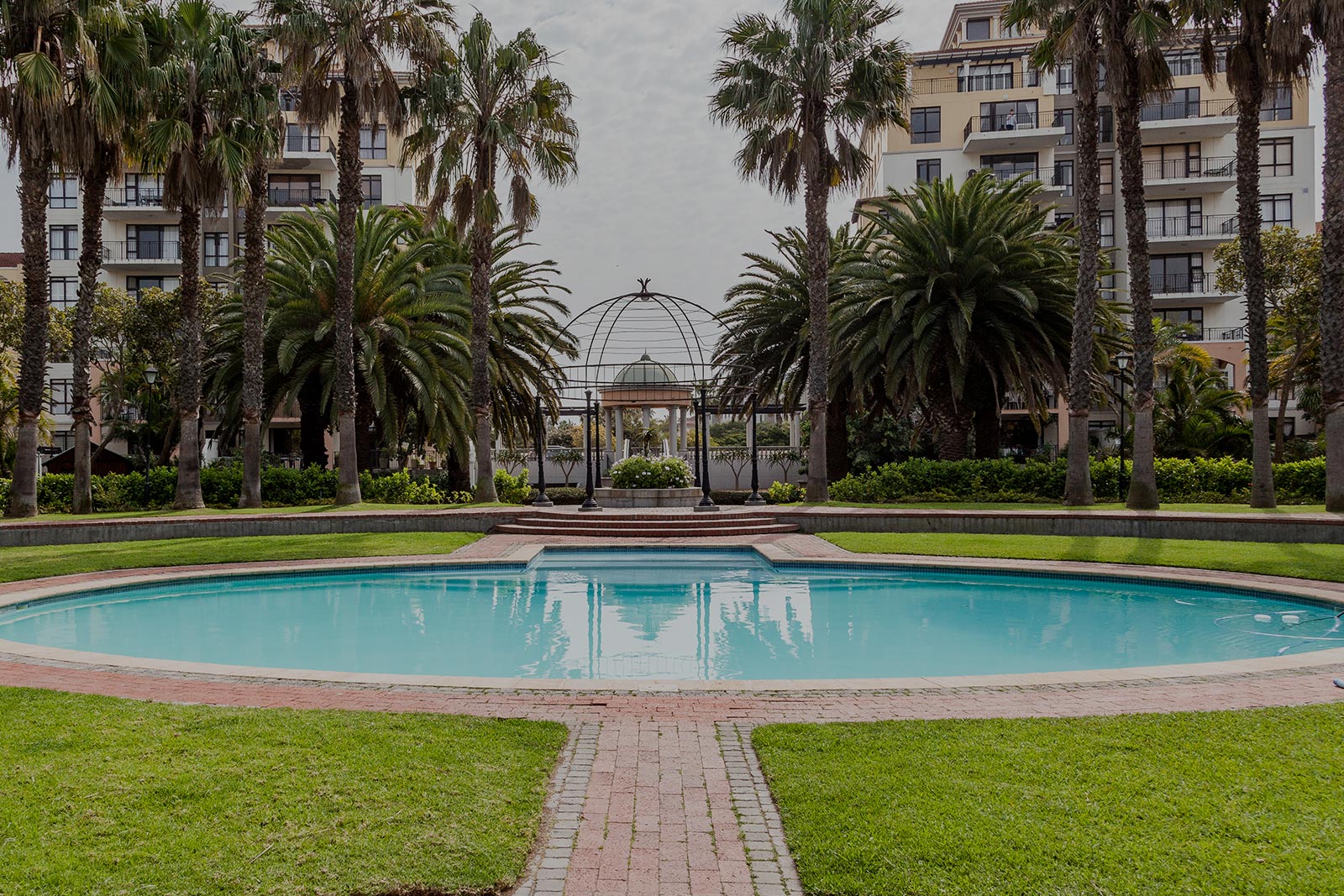 A circular swimming pool by Singer Group is surrounded by green grass, palm trees, and brick pathways, with a gazebo structure and apartment buildings in the background.