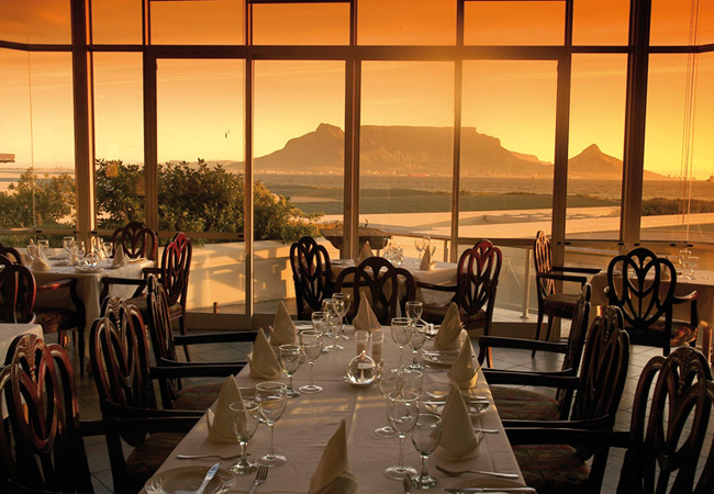 Elegant restaurant dining area with set tables and glass windows, overlooking a scenic sunset view of Table Mountain and the waterfront outside, brought to you by Singer Group.