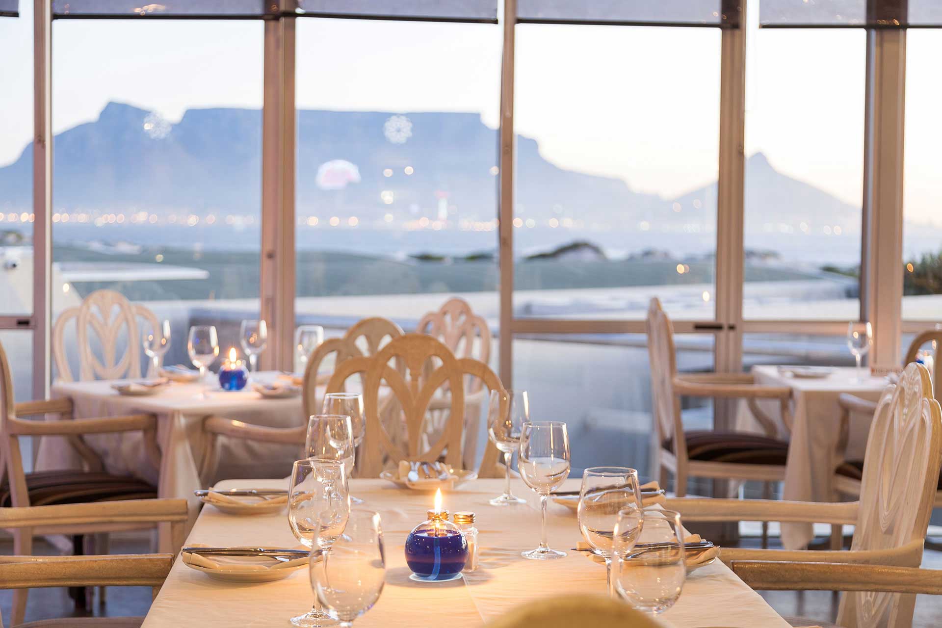 Elegant restaurant dining area with set tables, white tablecloths, glassware, and a blue candle centerpiece. Managed by Singer Group, the venue offers large windows revealing a scenic view of Table Mountain in the background.