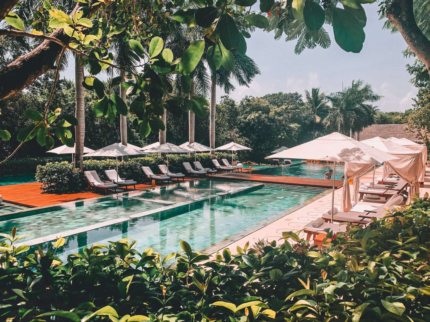 A luxurious outdoor pool by Singer Group is surrounded by tropical trees and greenery, with sun loungers and white umbrellas lining the poolside, and elegant white cabanas on the right under a sunny sky.