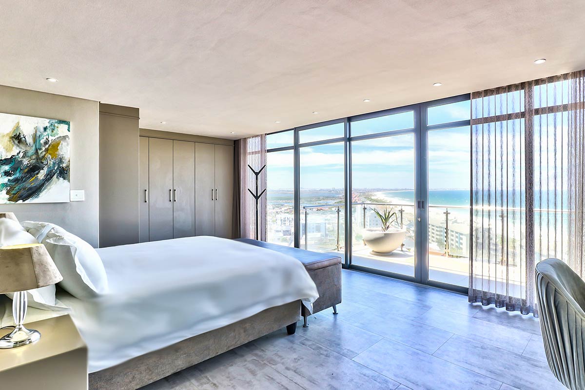 Modern bedroom with a large bed, white bedding, and minimalist decor by Singer Group. Floor-to-ceiling windows reveal a scenic ocean view and let in natural light, while sheer curtains frame the scene. A painting hangs on the wall.