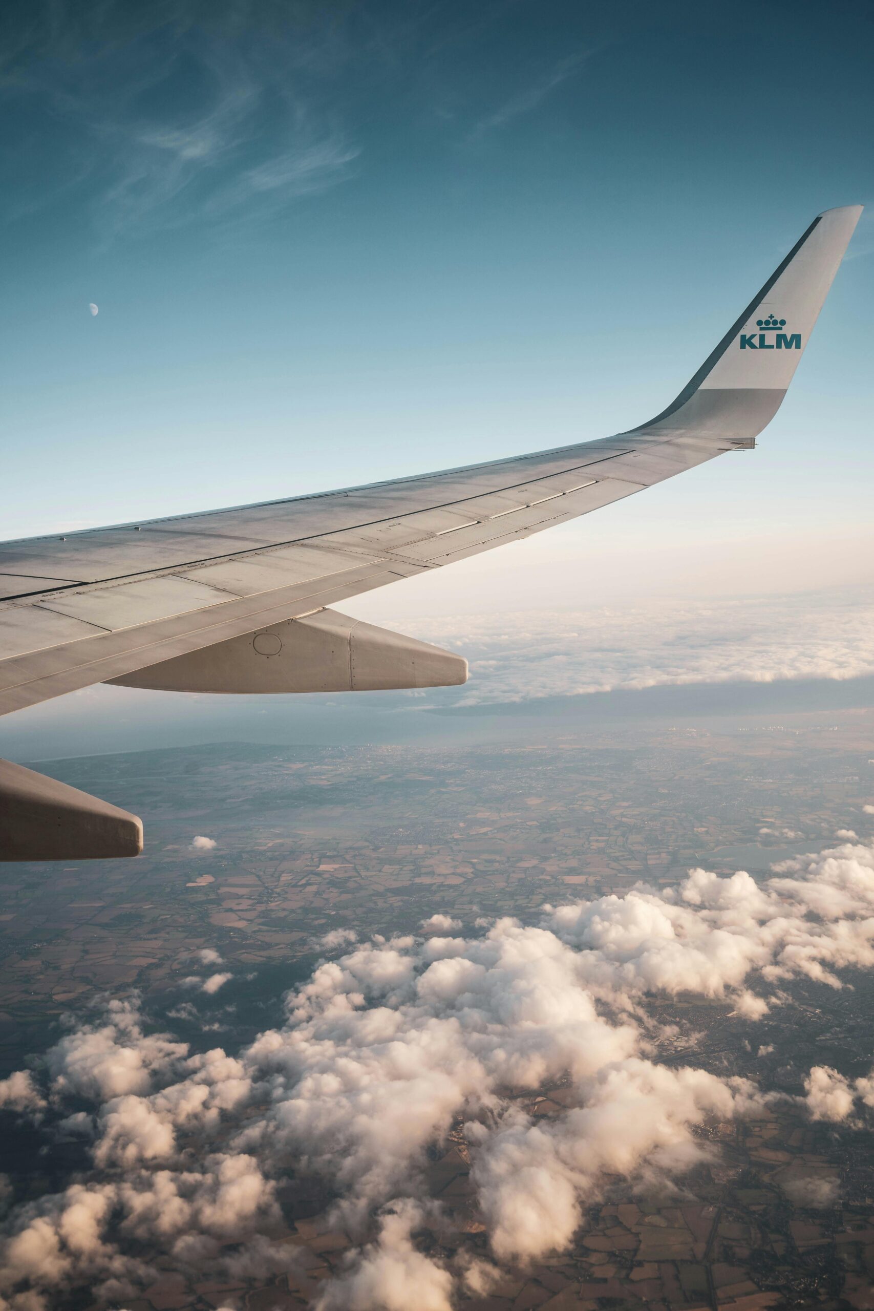 A view from an airplane window shows the KLM wing soaring above clouds and landscape below, with a clear blue sky in the background—a breathtaking scene on a Singer Group journey.
