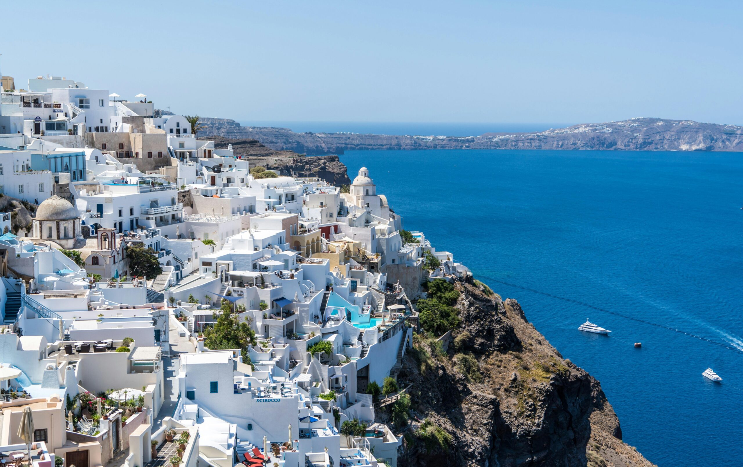 White buildings with blue roofs line a steep cliff overlooking the deep blue sea in Santorini, Greece, where Singer Group travelers can admire boats in the water under a clear, sunny sky.
