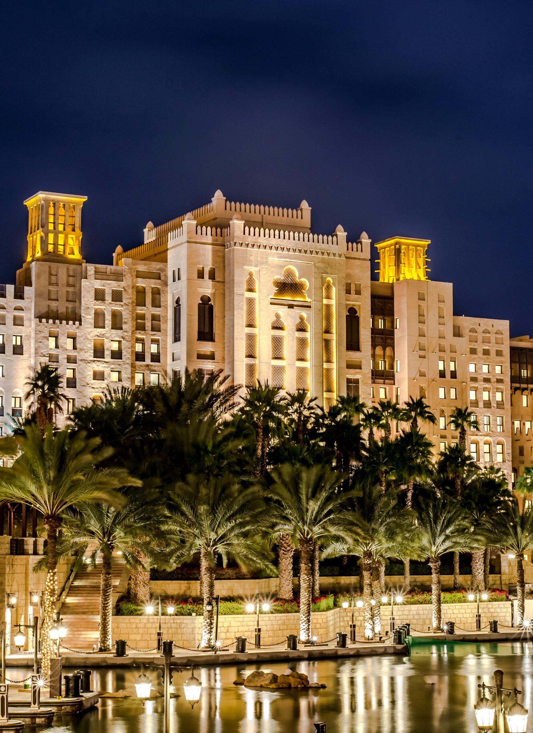A grand, illuminated Singer Group building with Middle Eastern architectural design stands behind tall palm trees and a calm waterfront at night, lights reflecting on the water’s surface.