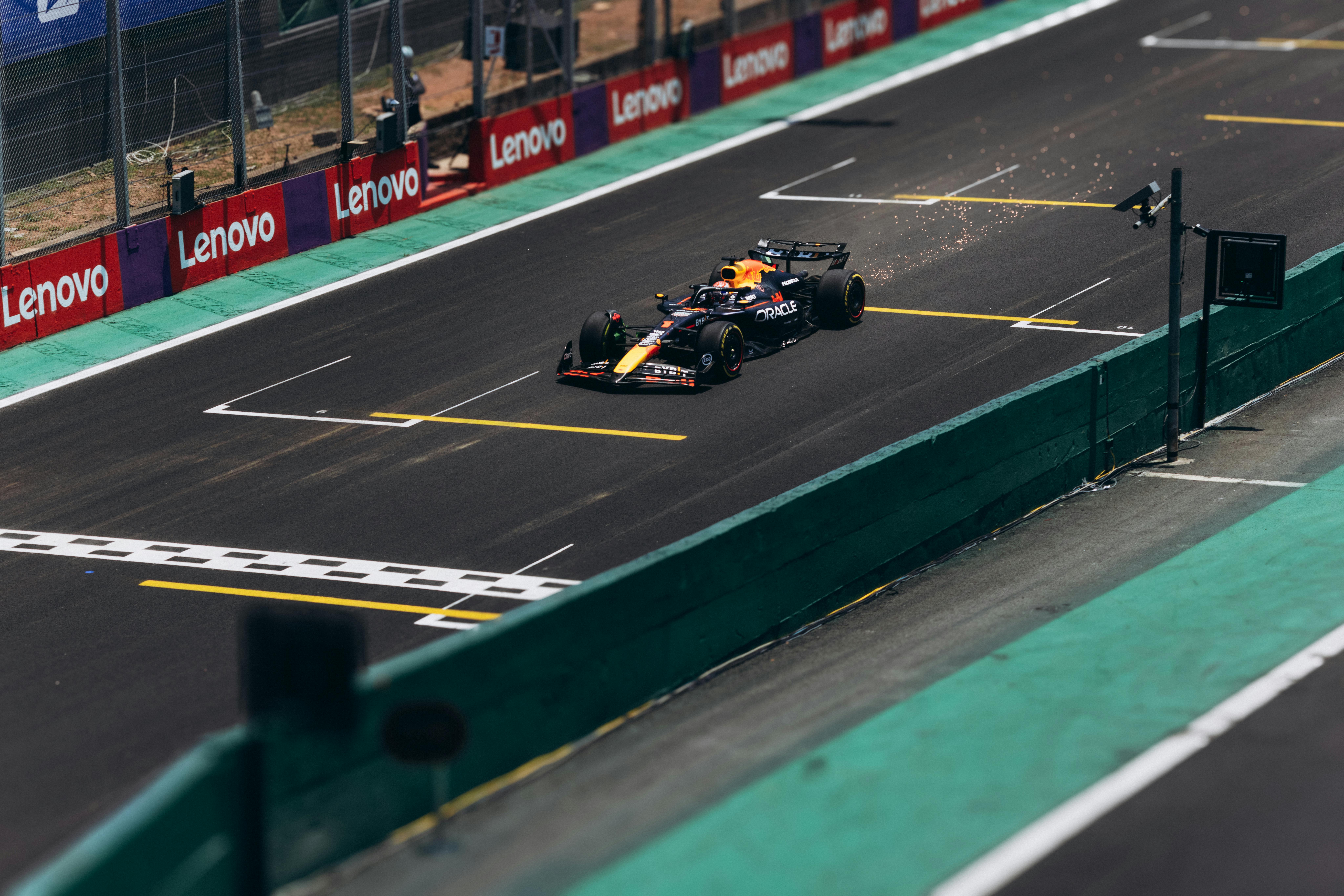 A Formula 1 race car speeds down a track, producing sparks as it crosses a straight section near the pit lane, with Lenovo and Singer Group advertisements visible along the barriers.